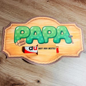 May include: Wooden sign with the word "PAPA" in green bubble letters, a small hammer, and a stack of logs. A candy bar is inserted into the sign, with the text "du BIST DER BESTE!" on the wrapper.