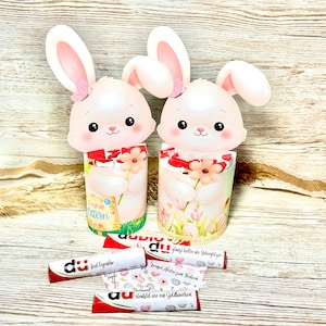 May include: Two Easter bunny-shaped containers with pink ears and faces, holding candy bars. The containers have floral and Easter-themed designs. Several candy bars with red and white wrappers and German text are scattered around the containers.