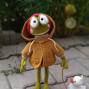 May include: A green crocheted frog wearing a brown hooded jacket and a red hat walks a white crocheted duck on a leash.