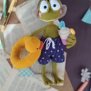 Può includere: Una rana di uncinetto verde che indossa un cappello marrone e pantaloncini viola con pois bianchi. La rana tiene un anello gonfiabile giallo e una tazza di uncinetto rosa e bianca con una cannuccia blu.