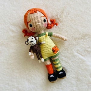 Puede incluir: Una mu&ntilde;eca de crochet con pelo naranja, que lleva un vestido amarillo y polainas a rayas verdes y naranjas, sosteniendo un mono de crochet.