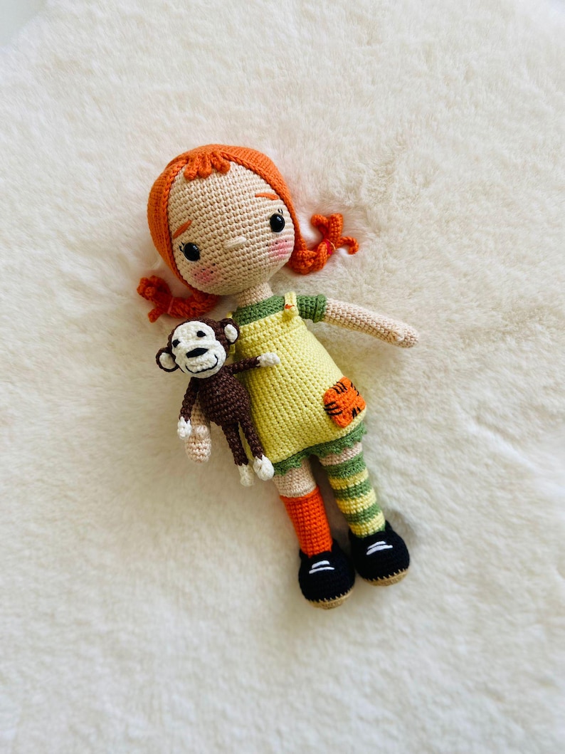 Puede incluir: Una mu&ntilde;eca de crochet con pelo naranja, que lleva un vestido amarillo y polainas a rayas verdes y naranjas, sosteniendo un mono de crochet.