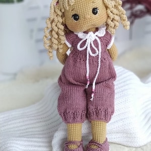 Op de afbeelding: Een gehaakte pop met krullend blond haar, die een roze en witte jurk en roze schoenen draagt. De pop zit op een wit deken.