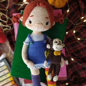 Puede incluir: Una mu&ntilde;eca de crochet con pelo rojo, un vestido azul y calcetines morados sostiene un mono de crochet marr&oacute;n con una pajarita amarilla. La mu&ntilde;eca est&aacute; sentada en una pila de libros con una tela de cuadros rojos y negros en el fondo.
