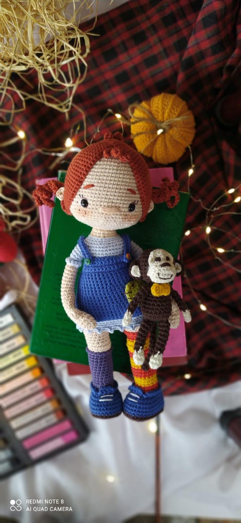 Puede incluir: Una mu&ntilde;eca de crochet con pelo rojo, un vestido azul y calcetines morados sostiene un mono de crochet marr&oacute;n con una pajarita amarilla. La mu&ntilde;eca est&aacute; sentada en una pila de libros con una tela de cuadros rojos y negros en el fondo.