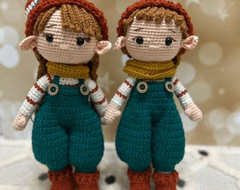 May include: Two crocheted elf dolls with brown hair, green overalls, and red boots. The dolls are wearing yellow scarves and have white and red striped shirts.