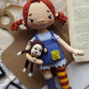 Puede incluir: Una mu&ntilde;eca de crochet con pelo rojo, un vestido azul y blanco y un mono de crochet en sus brazos. La mu&ntilde;eca lleva calcetines a rayas y zapatos azules. La mu&ntilde;eca est&aacute; sentada sobre una superficie de madera con una pancarta que dice "NR" en el fondo.