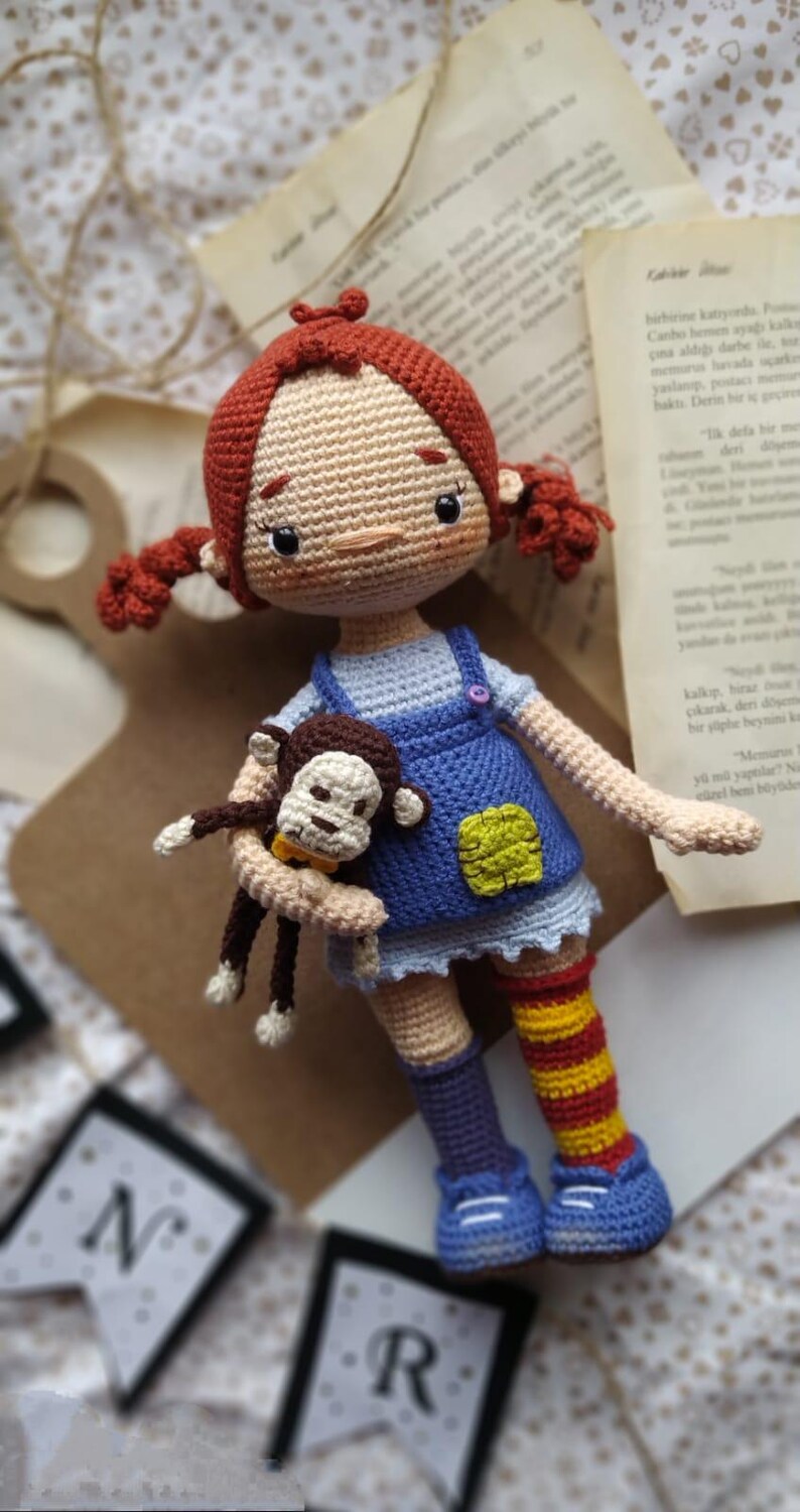 Puede incluir: Una mu&ntilde;eca de crochet con pelo rojo, un vestido azul y blanco y un mono de crochet en sus brazos. La mu&ntilde;eca lleva calcetines a rayas y zapatos azules. La mu&ntilde;eca est&aacute; sentada sobre una superficie de madera con una pancarta que dice "NR" en el fondo.