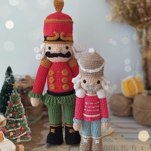 May include: Two crocheted nutcracker dolls, one large and one small. The larger doll is wearing a red and green uniform with a gold trim. The smaller doll is wearing a pink and blue uniform with a white trim. Both dolls have white yarn hair and black yarn eyes.