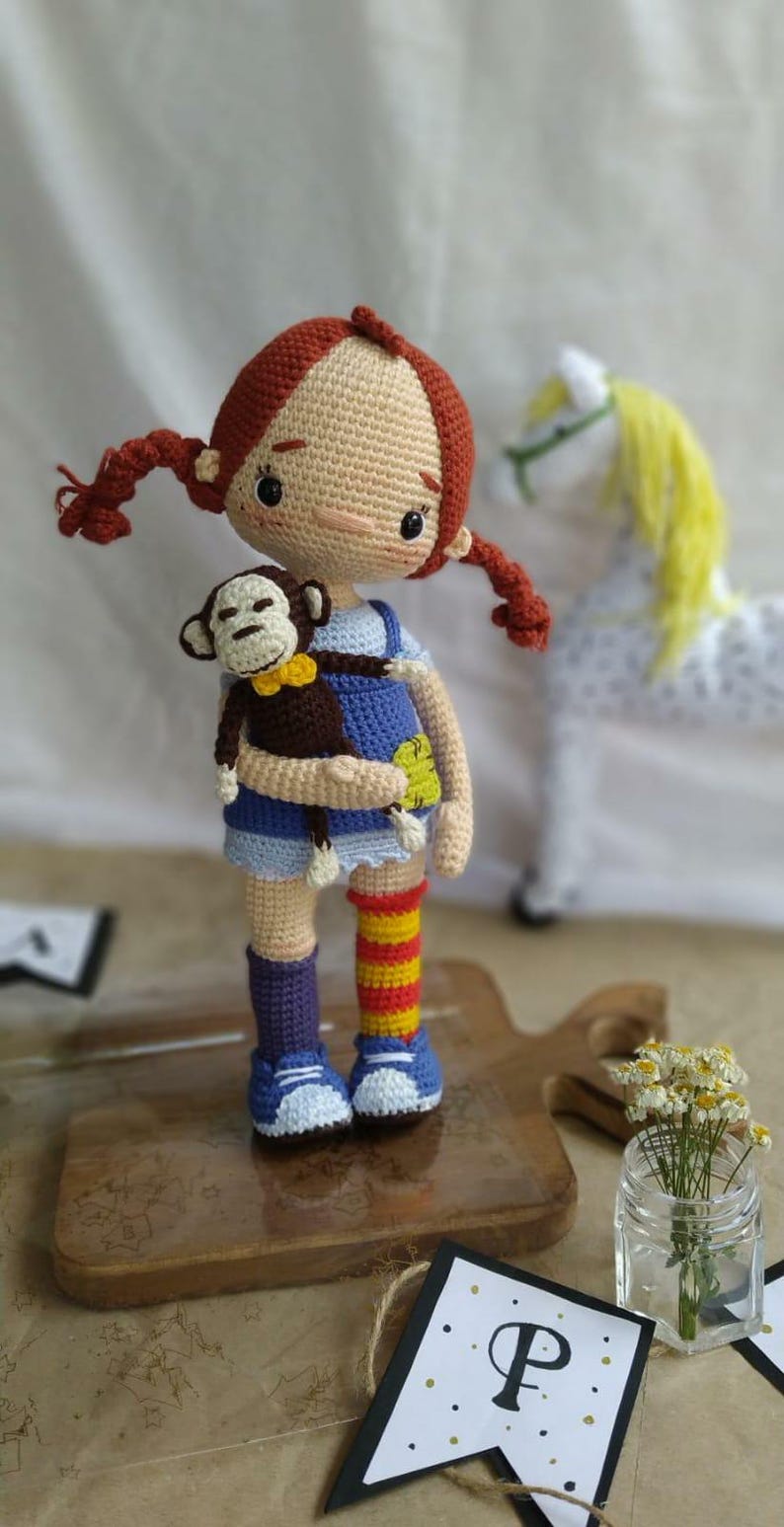 Puede incluir: Una mu&ntilde;eca de crochet con pelo rojo, vestida con un vestido azul y blanco y sosteniendo un mono de crochet marr&oacute;n. La mu&ntilde;eca est&aacute; de pie sobre una tabla de cortar de madera.