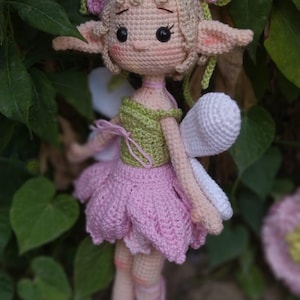May include: A handmade crochet fairy doll with a light skin tone, wearing a green top and a pink skirt. The doll has blonde hair, pink flower accents, and white wings. The doll is surrounded by green foliage.