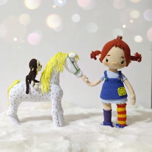 Puede incluir: Una mu&ntilde;eca de crochet con cabello rojo y un vestido azul sostiene la mano de un caballo de crochet blanco con una crin amarilla. Un peque&ntilde;o mono de crochet marr&oacute;n est&aacute; sentado en la espalda del caballo.