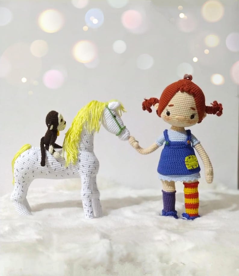 Puede incluir: Una mu&ntilde;eca de crochet con cabello rojo y un vestido azul sostiene la mano de un caballo de crochet blanco con una crin amarilla. Un peque&ntilde;o mono de crochet marr&oacute;n est&aacute; sentado en la espalda del caballo.