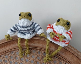 May include: Two knitted frog plushies wearing striped sweaters. One frog is wearing a grey and white striped sweater, and the other is wearing a red and white striped sweater. The frogs are sitting on a wicker chair.