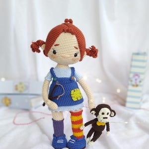 Puede incluir: Una mu&ntilde;eca de crochet con el pelo rojo en trenzas que lleva un vestido de peto azul y sostiene un mono de crochet marr&oacute;n. La mu&ntilde;eca lleva calcetines a rayas morados y naranjas y zapatos azules.