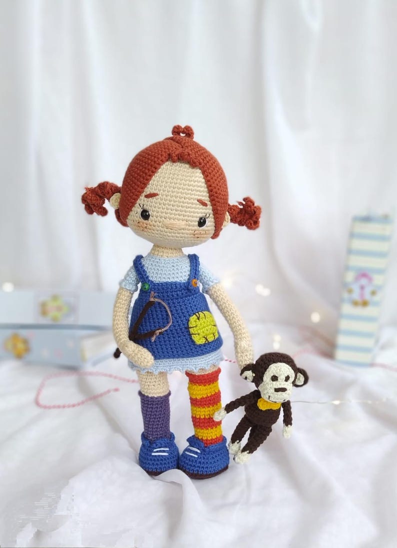 Puede incluir: Una mu&ntilde;eca de crochet con el pelo rojo en trenzas que lleva un vestido de peto azul y sostiene un mono de crochet marr&oacute;n. La mu&ntilde;eca lleva calcetines a rayas morados y naranjas y zapatos azules.