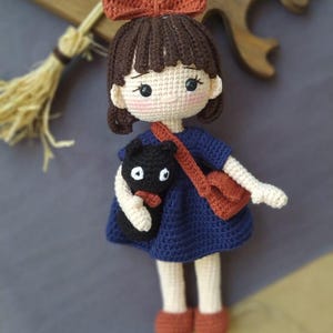 Könnte beinhalten: Gehäkelte Puppe eines Mädchens mit braunen Haaren, einer rostfarbenen Schleife und einem blauen Kleid. Sie hält eine schwarze Katze und eine rostfarbene Tasche. Die Puppe befindet sich auf einer grauen Oberfläche mit einem Holzbrett und einem Besen im Hintergrund.