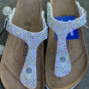 Puede incluir: Sandalias blancas de cuero con diseño de tira en T, adornadas con pedrería iridiscente. Las sandalias tienen una plantilla de corcho y un logotipo de Birkenstock.
