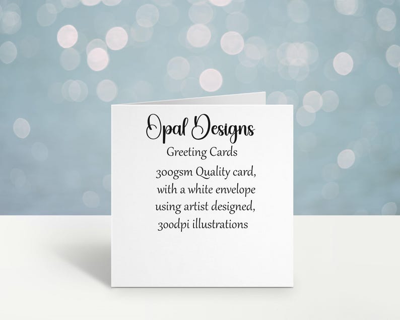 May include: A white greeting card from Opal Designs, with text that reads "Greeting Cards, 300gsm Quality card, with a white envelope using artist designed, 300dpi illustrations." The card is on a white surface with a blurred blue background.