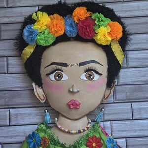Puede incluir: Un adorno de pared decorativo que representa un rostro estilizado con cabello oscuro y una colorida corona floral. La figura lleva una blusa verde bordada con detalles florales vibrantes, un collar de cuentas y pendientes de borlas azules. El fondo es un patr&oacute;n de ladrillos.