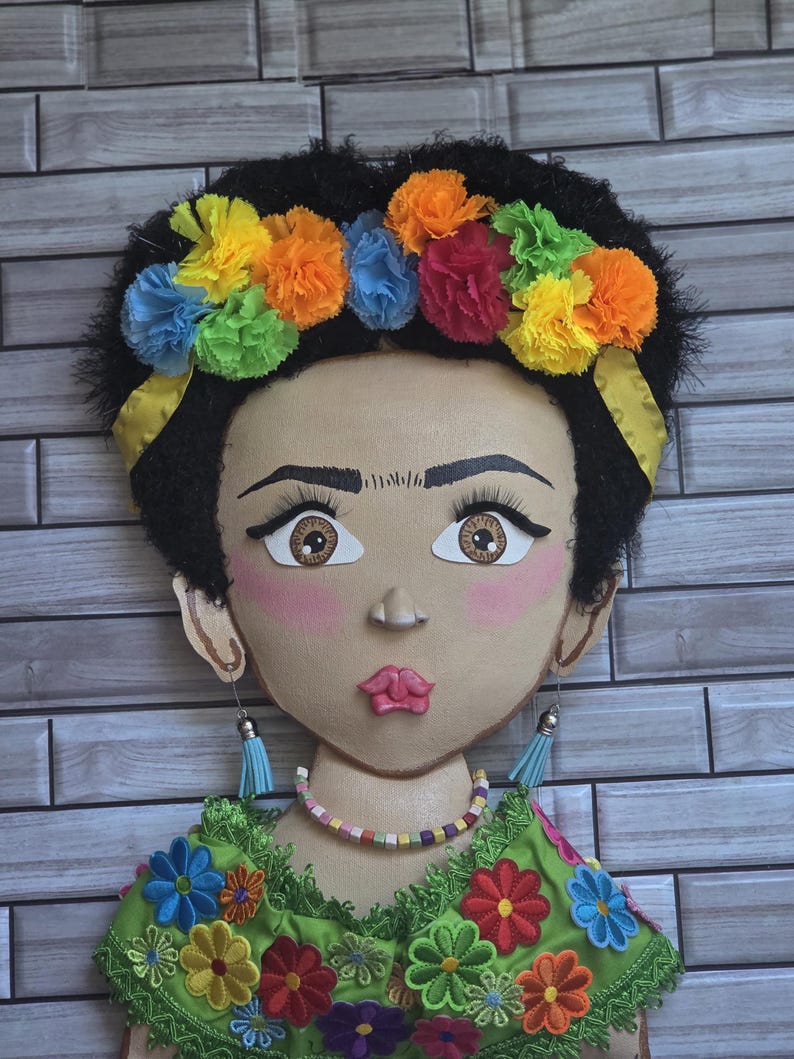 Puede incluir: Un adorno de pared decorativo que representa un rostro estilizado con cabello oscuro y una colorida corona floral. La figura lleva una blusa verde bordada con detalles florales vibrantes, un collar de cuentas y pendientes de borlas azules. El fondo es un patr&oacute;n de ladrillos.