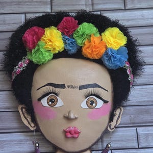 Puede incluir: Decoraci&oacute;n mural que representa un rostro estilizado con cabello oscuro y una colorida corona floral. El rostro tiene rasgos pintados, incluyendo ojos grandes, rubor rosa y un collar. Los pendientes son borlas moradas.