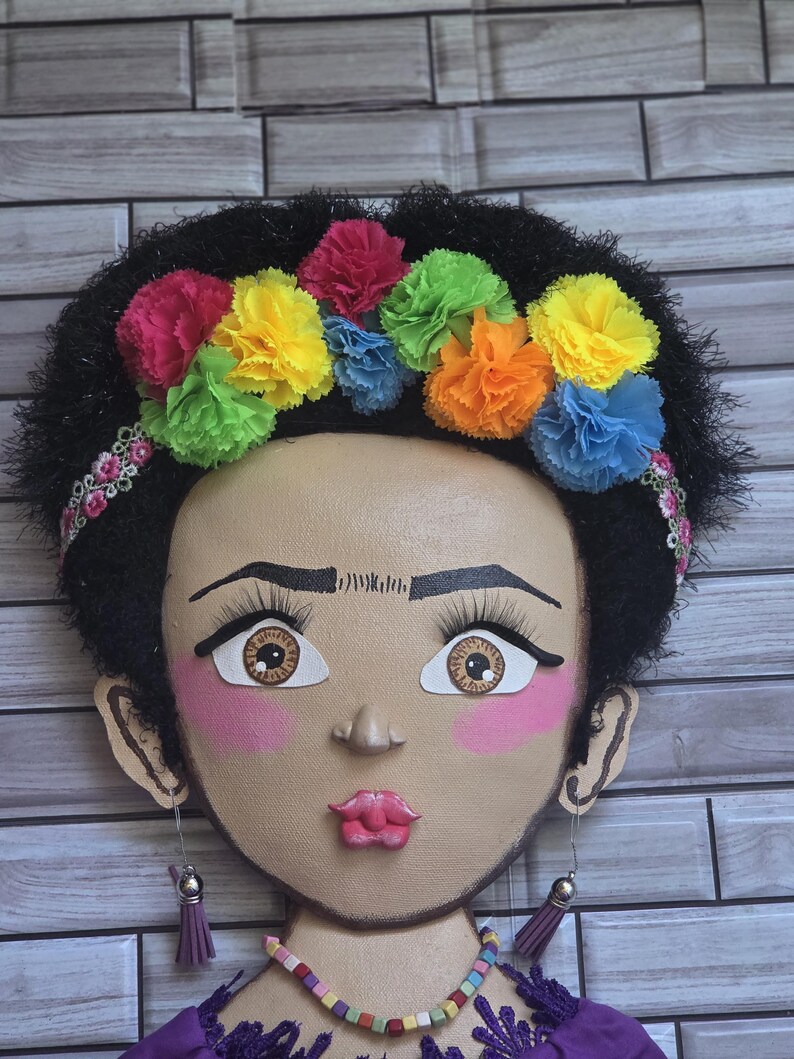 Puede incluir: Decoraci&oacute;n mural que representa un rostro estilizado con cabello oscuro y una colorida corona floral. El rostro tiene rasgos pintados, incluyendo ojos grandes, rubor rosa y un collar. Los pendientes son borlas moradas.