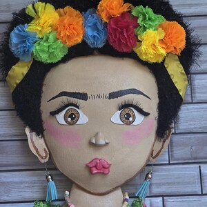 Puede incluir: Un adorno de pared decorativo que representa una cara pintada adornada con una corona floral. La cara tiene cabello oscuro, ojos grandes y rubor rosa. La corona est&aacute; hecha de flores de papel de colores. Los pendientes son borlas azules. El collar est&aacute; hecho de cuentas de colores.