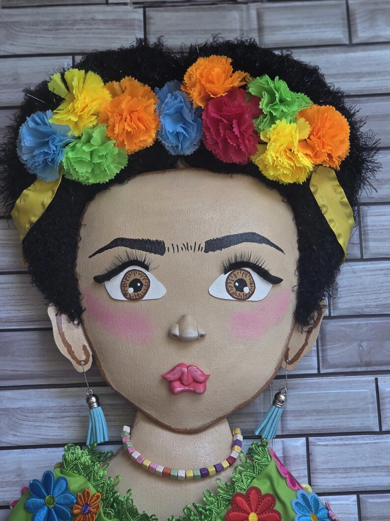 Puede incluir: Un adorno de pared decorativo que representa una cara pintada adornada con una corona floral. La cara tiene cabello oscuro, ojos grandes y rubor rosa. La corona est&aacute; hecha de flores de papel de colores. Los pendientes son borlas azules. El collar est&aacute; hecho de cuentas de colores.