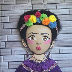 Puede incluir: Un adorno de pared artesanal que representa un rostro estilizado con cabello oscuro y una corona floral colorida. El rostro tiene mejillas rosadas, ojos grandes y un vestido morado con ribete de encaje.