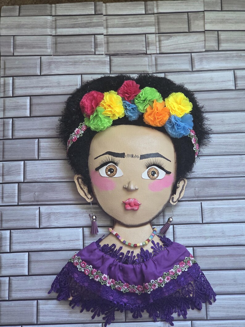 Puede incluir: Un adorno de pared artesanal que representa un rostro estilizado con cabello oscuro y una corona floral colorida. El rostro tiene mejillas rosadas, ojos grandes y un vestido morado con ribete de encaje.
