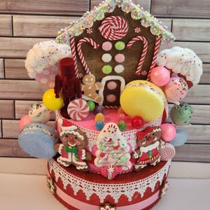 May include: A two-tiered Christmas cake decorated with gingerbread houses, candy canes, gumdrops, and gingerbread people. The cake is topped with a gingerbread house and a white and pink candy cane.