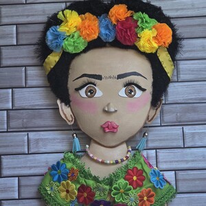 Puede incluir: Decoraci&oacute;n mural que presenta un retrato estilizado de una mujer con cabello oscuro y una corona floral colorida. El retrato tiene ojos grandes, mejillas rosadas y una blusa verde bordada con flores. Los aretes son azules.