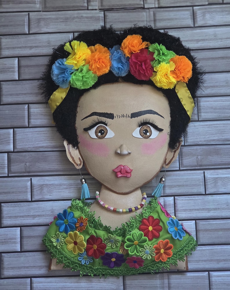 Puede incluir: Decoraci&oacute;n mural que presenta un retrato estilizado de una mujer con cabello oscuro y una corona floral colorida. El retrato tiene ojos grandes, mejillas rosadas y una blusa verde bordada con flores. Los aretes son azules.