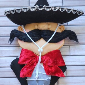 Puede incluir: Adorno de pared decorativo que representa una cara estilizada con un sombrero negro ribeteado en plata, un gran bigote negro y una pajarita de terciopelo rojo. La cara tiene un tono de piel claro y lleva una chaqueta negra.