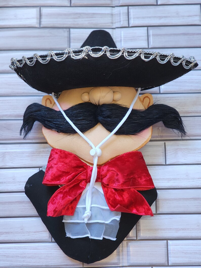 Puede incluir: Adorno de pared decorativo que representa una cara estilizada con un sombrero negro ribeteado en plata, un gran bigote negro y una pajarita de terciopelo rojo. La cara tiene un tono de piel claro y lleva una chaqueta negra.