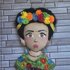 Puede incluir: Un adorno de pared decorativo que presenta un retrato estilizado de una mujer con cabello oscuro y una corona de flores. La cara est&aacute; pintada con mejillas rosadas y labios llenos. Lleva una blusa verde adornada con flores bordadas coloridas y pendientes colgantes.
