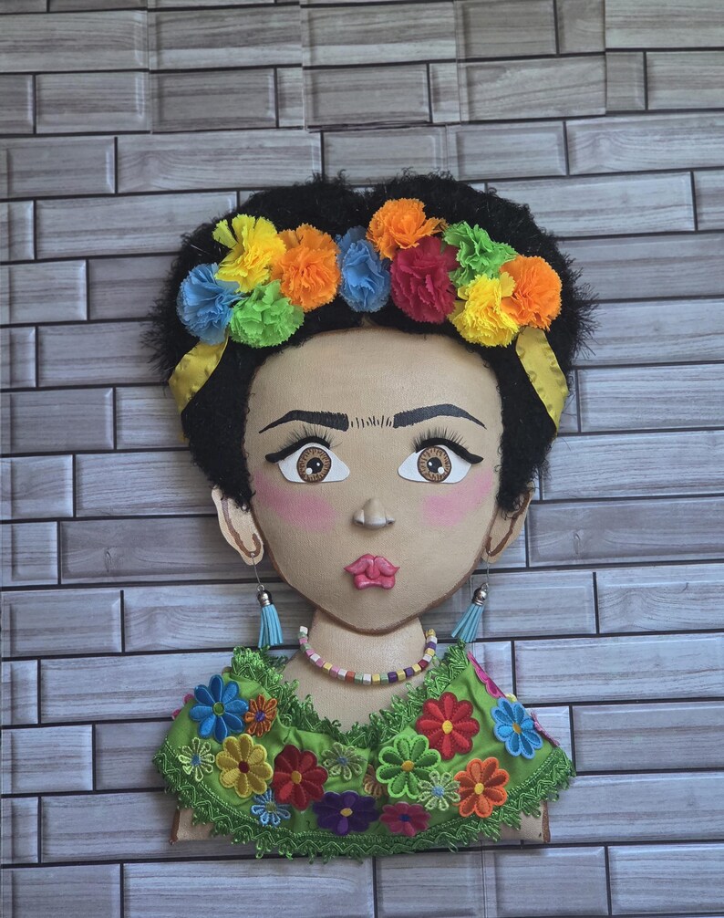 Puede incluir: Un adorno de pared decorativo que presenta un retrato estilizado de una mujer con cabello oscuro y una corona de flores. La cara est&aacute; pintada con mejillas rosadas y labios llenos. Lleva una blusa verde adornada con flores bordadas coloridas y pendientes colgantes.