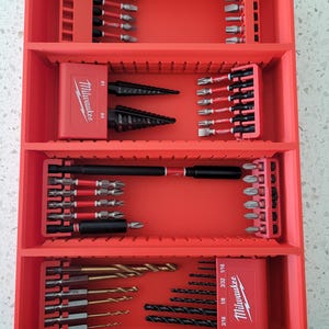 May include: A red tool organizer filled with various drill bits and accessories. The organizer has multiple compartments holding different sizes and types of bits, including step bits and drill bits. The Milwaukee logo is visible on a red case.