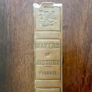 May include: A vintage book titled "Makers of History" with the name "Pyrrhus" printed on the spine. The book cover is a light brown color with a gold-colored design.