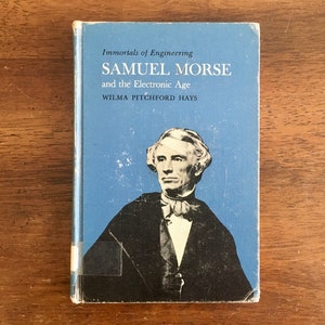 May include: A vintage book titled "Samuel Morse and the Electronic Age" with a blue cover and a portrait of Samuel Morse. The book is part of the "Immortals of Engineering" series. The book is on a wooden surface.