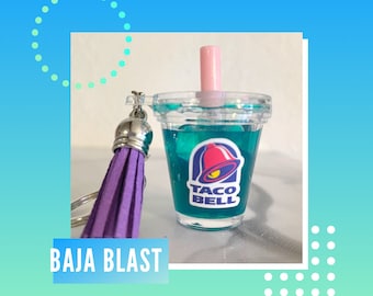 May include: A clear, miniature Baja Blast drink charm with a pink straw and a purple tassel keychain. The Taco Bell logo is visible on the front of the cup. The text "BAJA BLAST" is displayed on a white and blue rectangle.