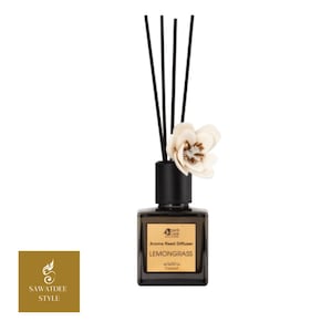 Könnte beinhalten: Aroma-Reed-Diffusor mit schwarzen Stäbchen und weißer Blütenakzent. Die quadratische, dunkle Glasflasche trägt die Aufschrift "Lemongrass". Das Produkt stammt von "Sawasdee Style".
