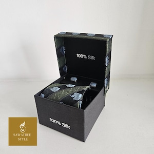 Puede incluir: Un juego de corbata y gemelos de seda negra con un estampado de elefantes, presentado en una caja negra. La corbata presenta un patrón repetido de elefantes en rayas azul claro y verde. La caja está etiquetada como "100% Seda".