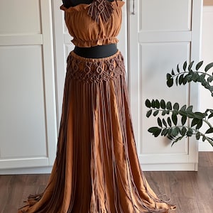 May include: A two-piece set consisting of a rust-colored off-the-shoulder crop top and a matching long skirt. Both pieces feature dark brown macrame detailing with intricate knotted patterns and long, hanging fringe elements throughout.