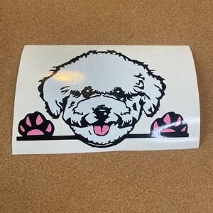 Op de afbeelding: Een wit en zwarte vinyl sticker met een cartoon hondengezicht met een roze tong en poten. De hond lacht en kijkt over een zwarte lijn, met roze pootafdrukken aan weerszijden.