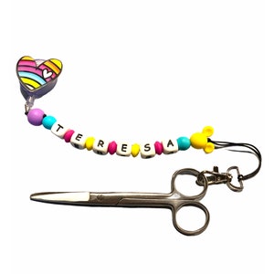 May include: A colourful beaded keychain with the name "TERESA" spelled out in white beads. The keychain has a rainbow heart charm and a yellow Mickey Mouse charm. A pair of silver scissors are also included.