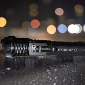 May include: A black flashlight with a textured grip and a metal clip. The text "NAME HERE" is printed on the side. The flashlight is cylindrical and has a ribbed bezel. The background is blurred with bokeh lights.
