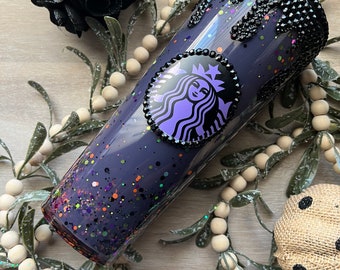 May include: A purple and black tumbler with a glittery finish and a black rhinestone rim. The tumbler has a Starbucks logo with a purple mermaid and a black crown.