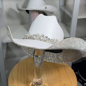May include: White cowboy hat with a wide brim, heavily embellished with sparkling rhinestones. The hat features a decorative rhinestone band and is displayed on a clear, faceted stand. Another white cowboy hat is visible in the background.